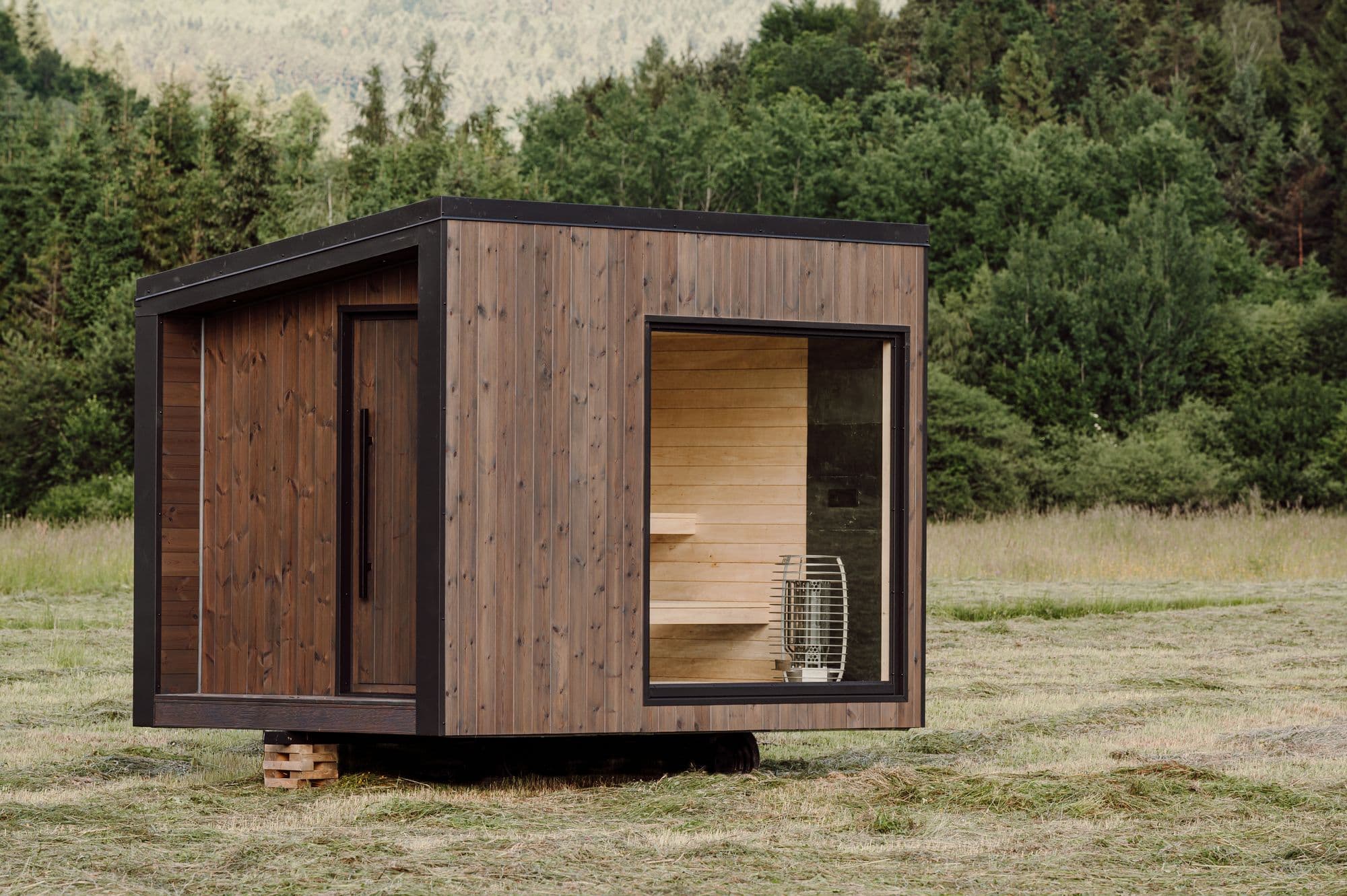 Sauna Hansei na skoszonej letniej łące, okno panoramiczne odsłania osikowe ławy i piec HUUM