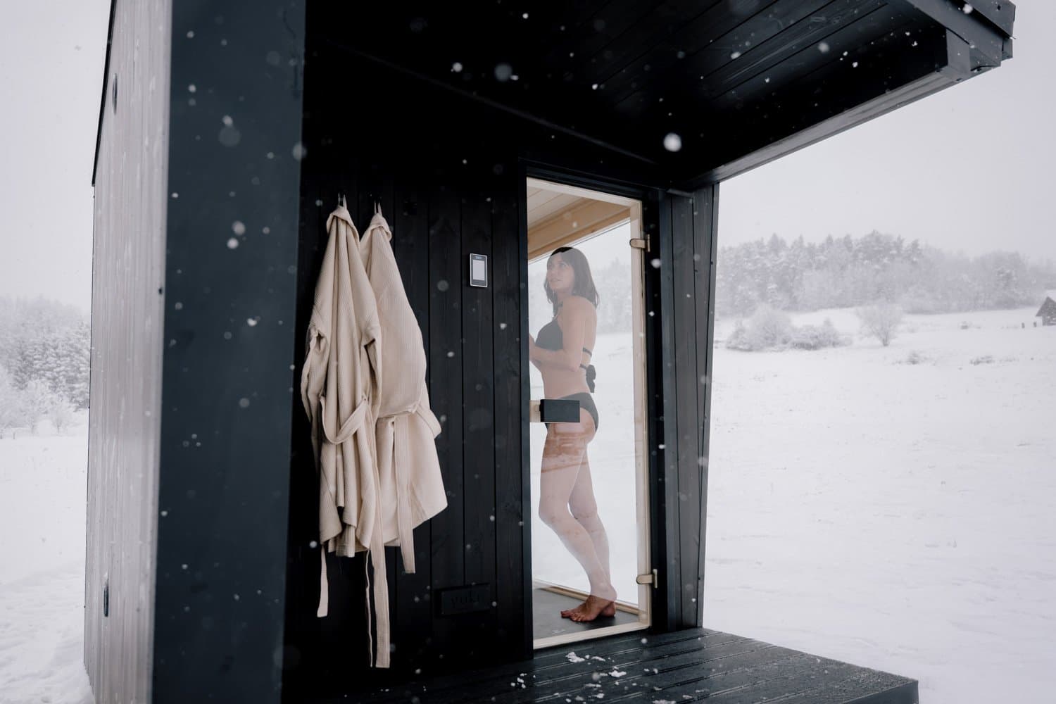 Ein Gast tritt bei Schneefall auf die überdachte Veranda der Sauna Ikigai 5, Bademäntel hängen an der Tür