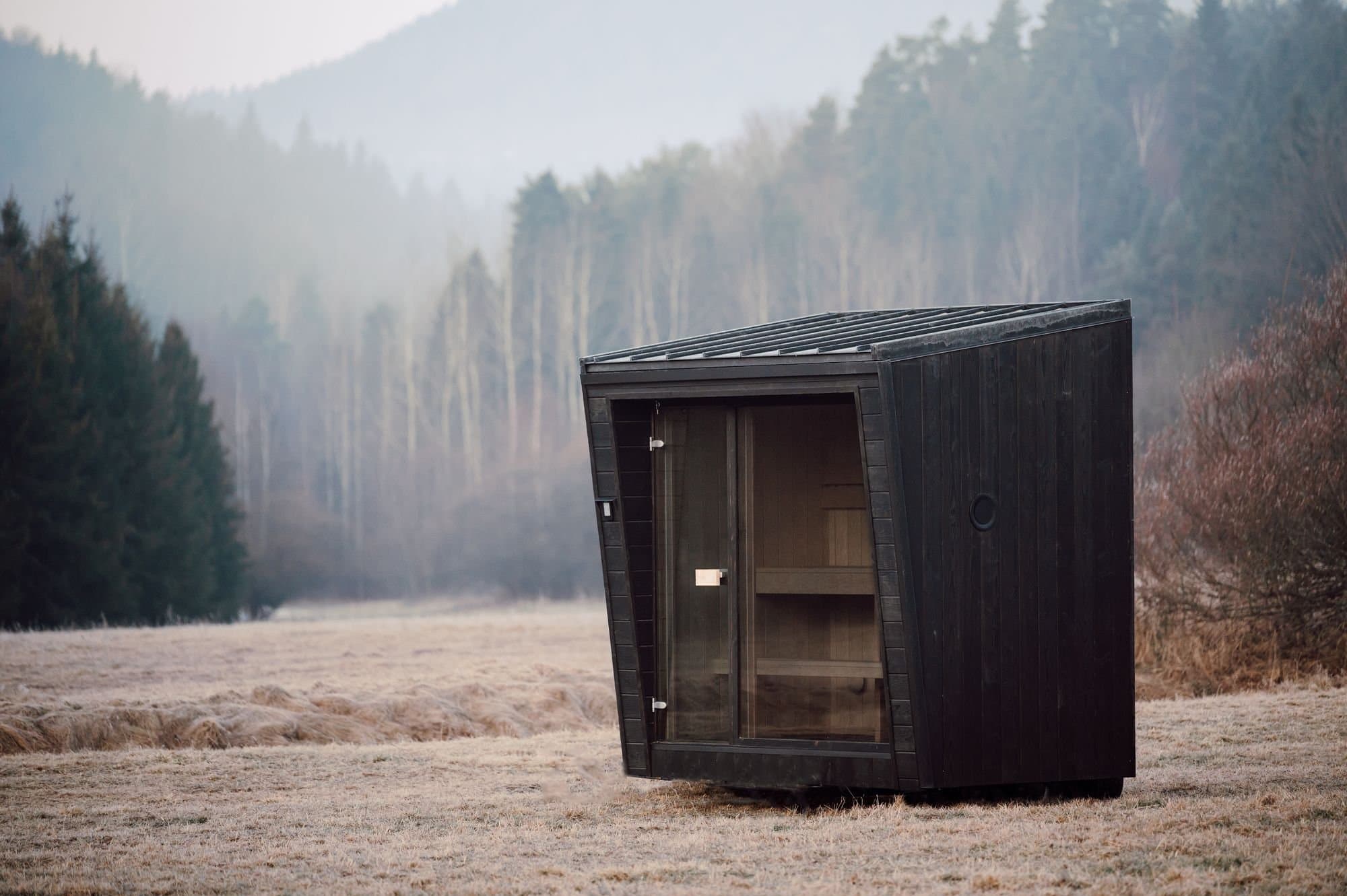 Sauna Kaizen auf einer bereiften Wiese unter nebligen Waldhügeln, Bänke durch die Glasfront sichtbar