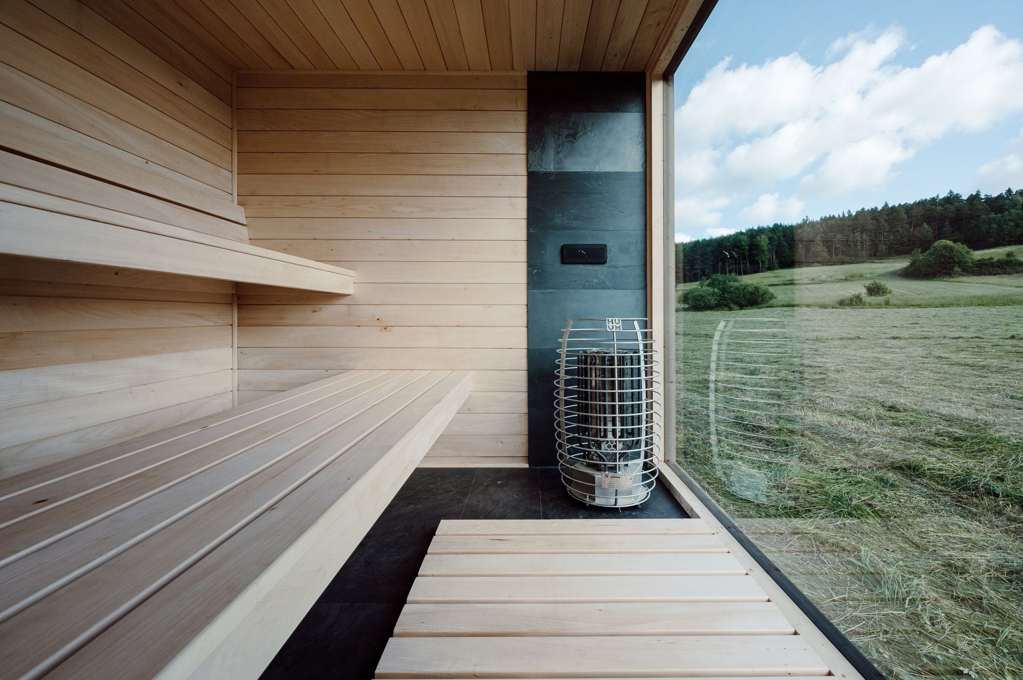 Innenraum einer Yuki Sauna Hansei mit zwei Bankebenen aus Lindenholz, einem Huum-Ofen vor einer dunklen Schieferwand und einem Fenster mit Blick auf eine Wiese